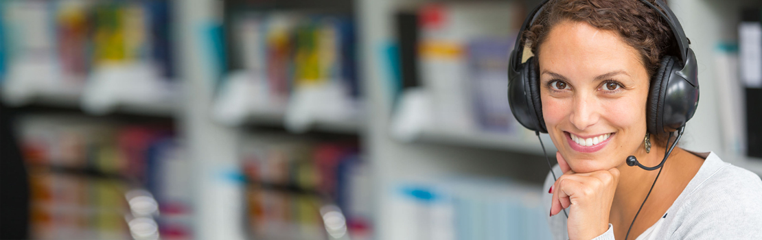 Imagebild für TestDaF Lehrkraefte; eine junge Frau mit Headset blickt in die Kamera, im Hintergrund sieht man eine Bücherwand